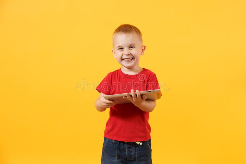 Little cute kid baby boy 3-4 years old in red t-shirt holding in hand tablet pc computer isolated on yellow background. Kids childhood lifestyle concept. Problem of children and gadgets. Copy space. Little cute kid baby boy 3-4 years old in red t-shirt holding in hand tablet pc computer isolated on yellow background. Kids childhood lifestyle concept. Problem of children and gadgets. Copy space